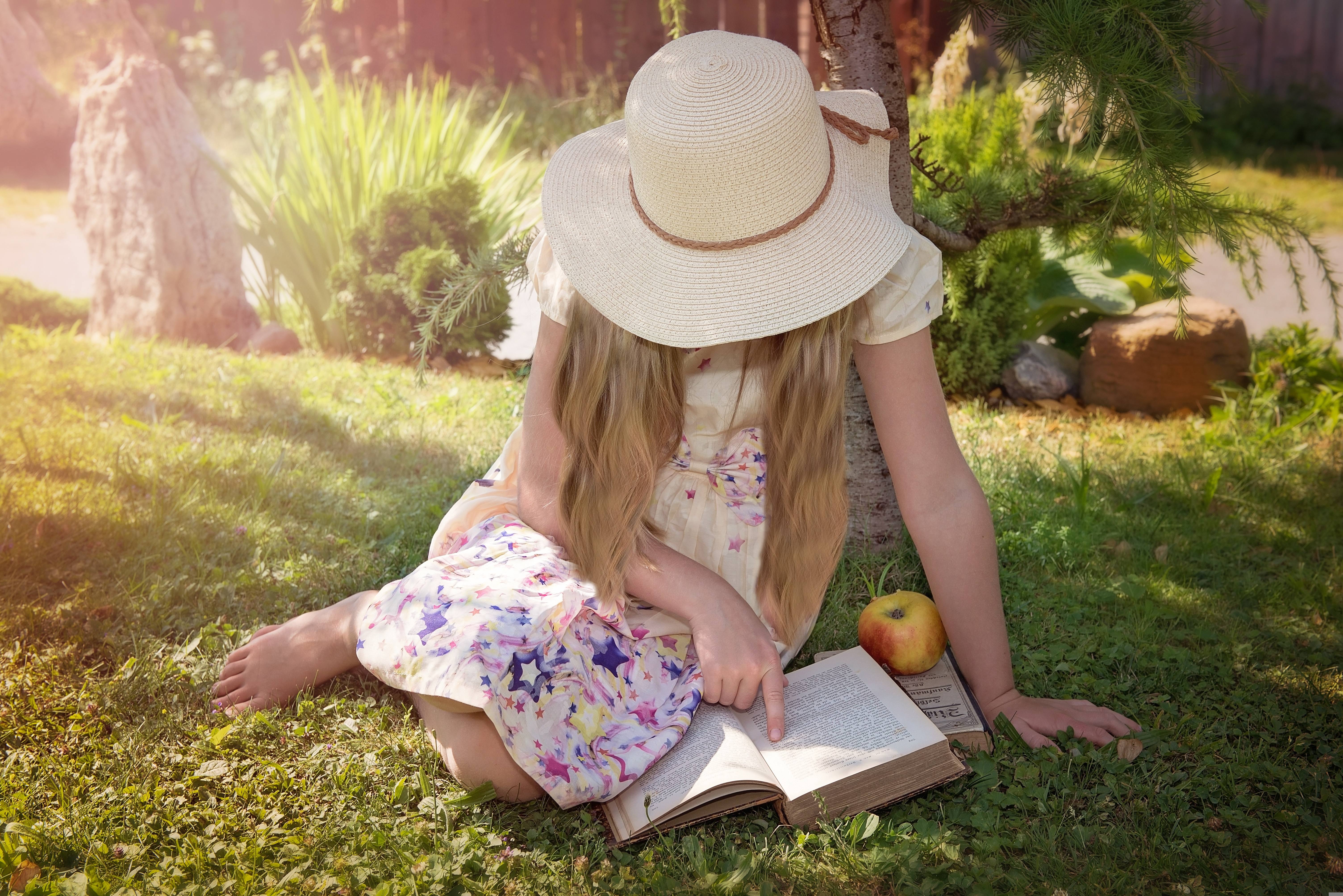 little girl in the yard with a sun hat on, reading a book