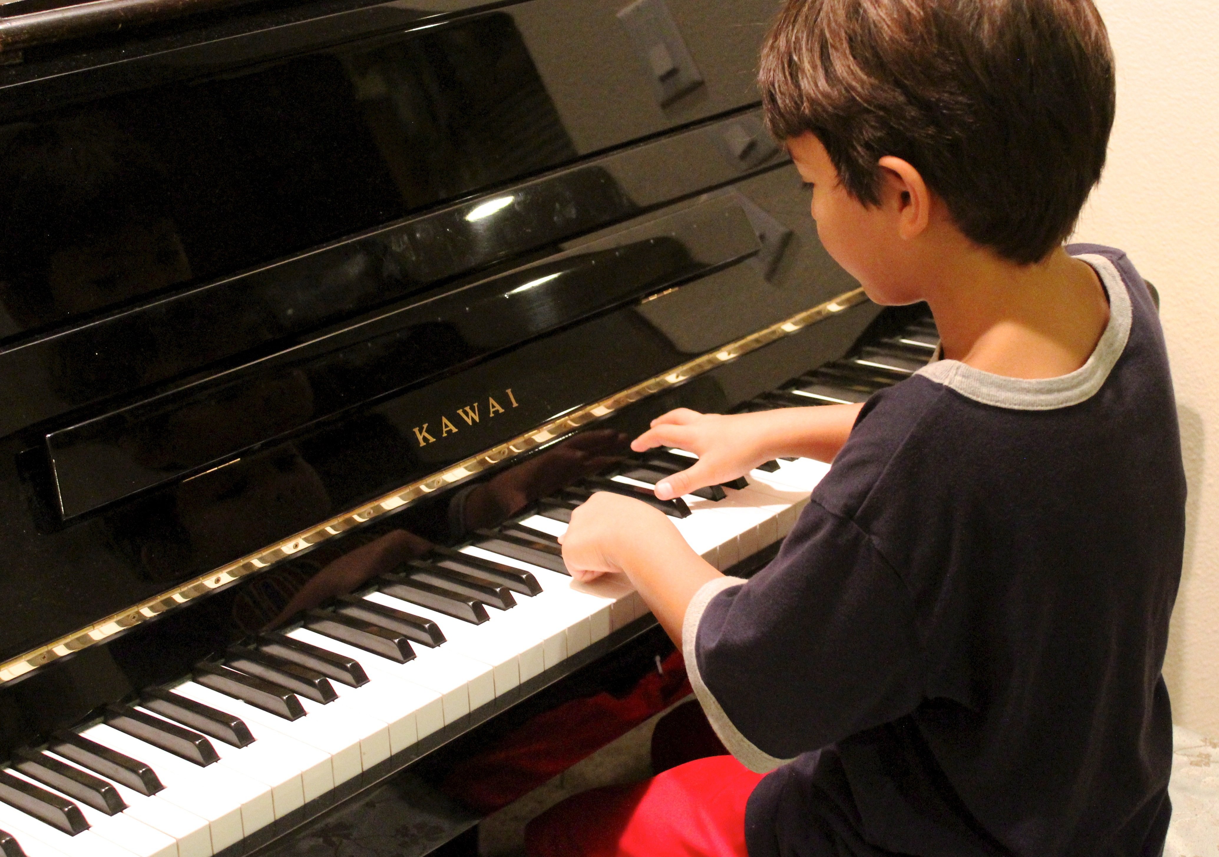 over-the-shoulder view of a little boy sitting on a piano bench, playing a Kawai piano